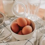 Close-up of fresh eggs in a bowl with kitchen utensils on a striped cloth for cooking preparation.